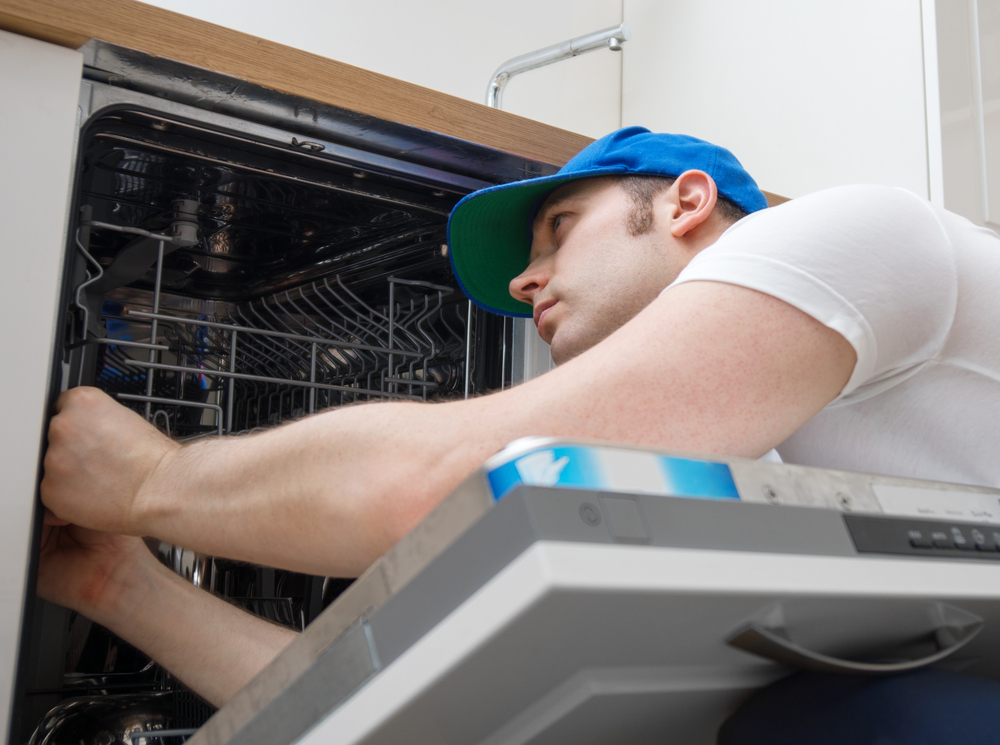 Professional handyman in overalls repairing domestic dishwasher in the kitchen.